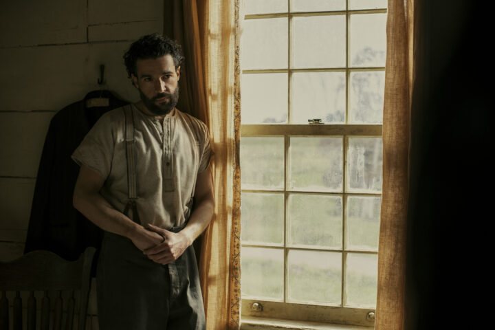 "A man with a beard stands pensively near a sunlit window in a rustic room, dressed in an old-fashioned shirt and trousers. The environment suggests a historical or rural setting with muted colors and a calm mood."
