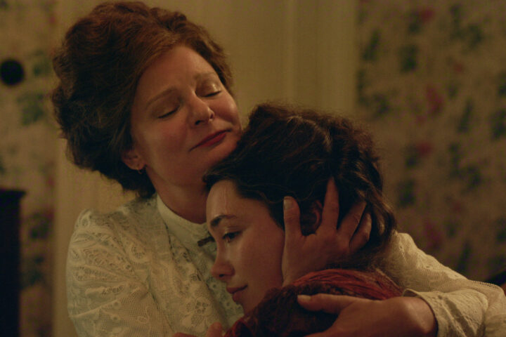 "Two women in vintage clothing share an emotional embrace in a warmly lit, floral-wallpapered room, suggesting comfort and support in a historical or period setting."